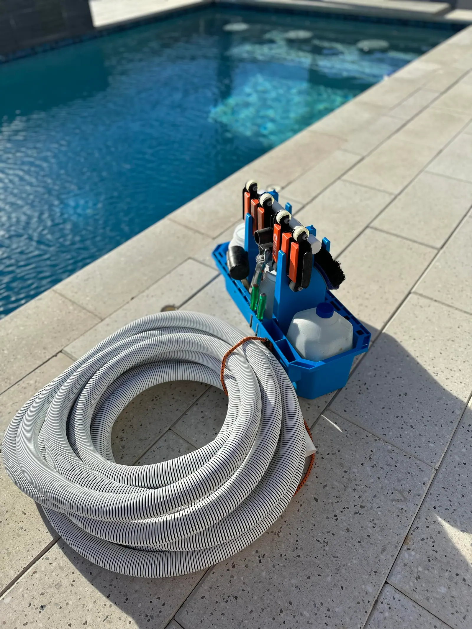 A coiled white vacuum hose and a blue caddy of pool tools on the pool deck