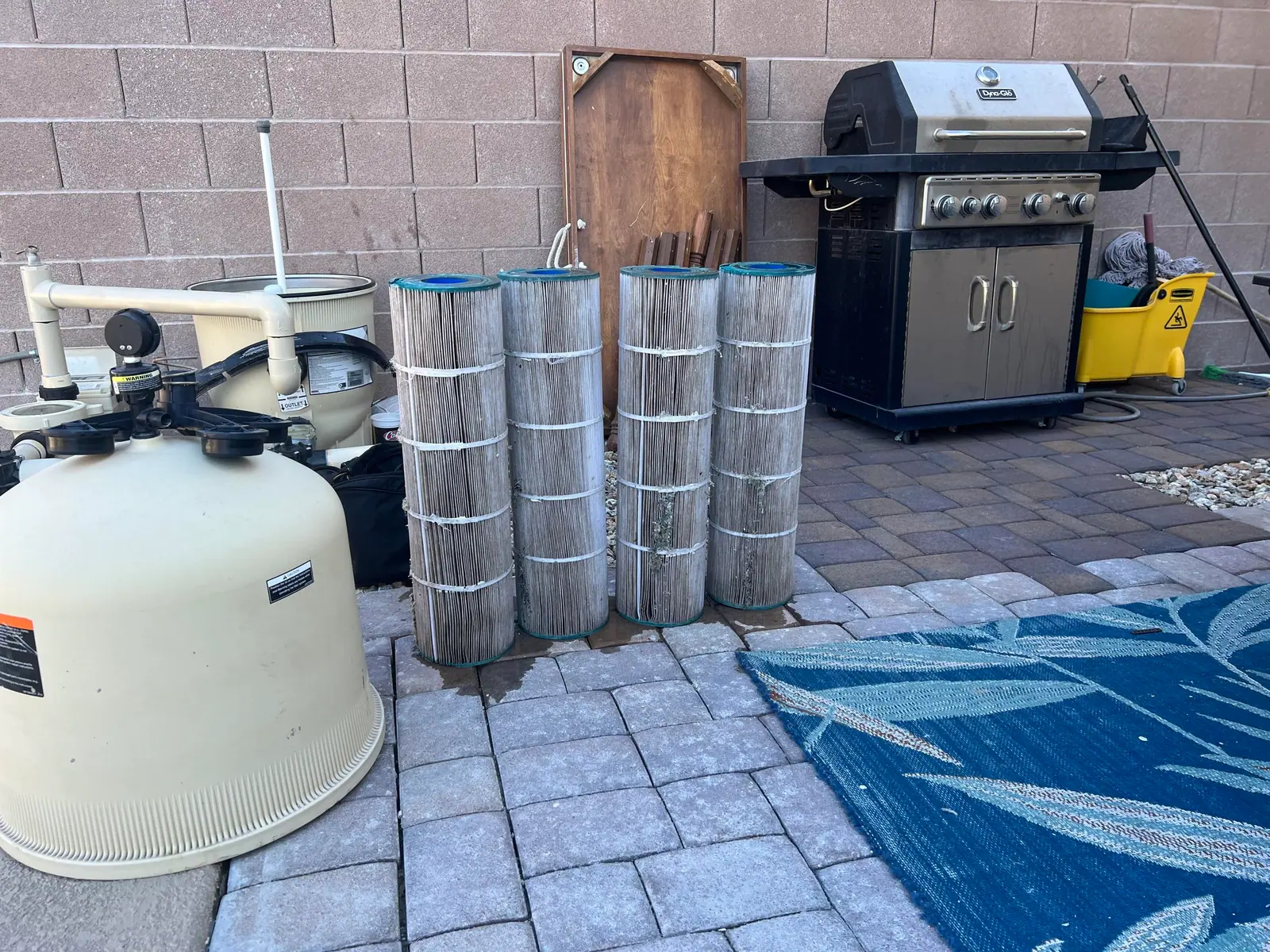 A pool pump, filter tank and cartridges on a paved patio beside a grill